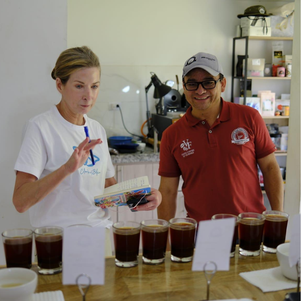 Ilse Geyskens and Wilton Benitez at a coffee tasting event at granja paraiso 92 with multiple cups of coffee arranged in front of them, along with coffee-related equipment and materials visible on the table.